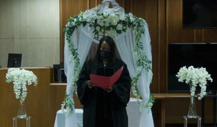 Black woman in judge's robe wearing mask standing in front of white wedding decorations and flowers reds from a red file folder