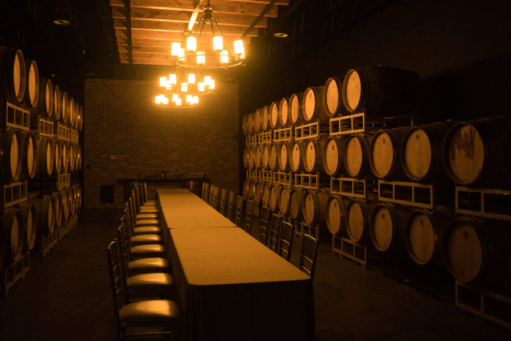 Inside of a dark cellar-like room with long table and lots of barrels of wine