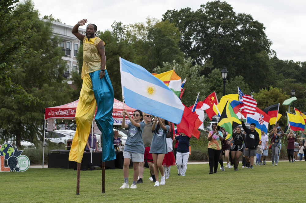 A group of people carrying various flags walk behind a Black man on stilts outside