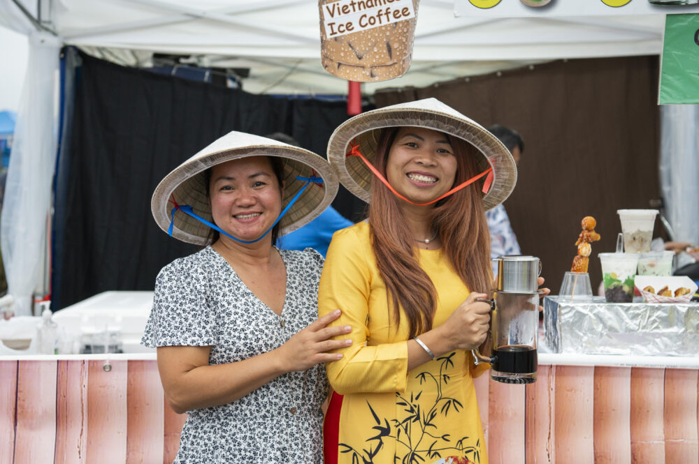 Two Asian women in colorful clothes and hats stand outside a coffee stand at a festival