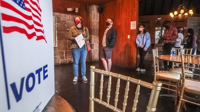 three people stand in line inside a polling place next to a voting booth wearing casual clothing