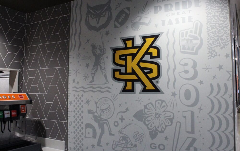 A gray and white wall with college logos of an owl, football and KSU