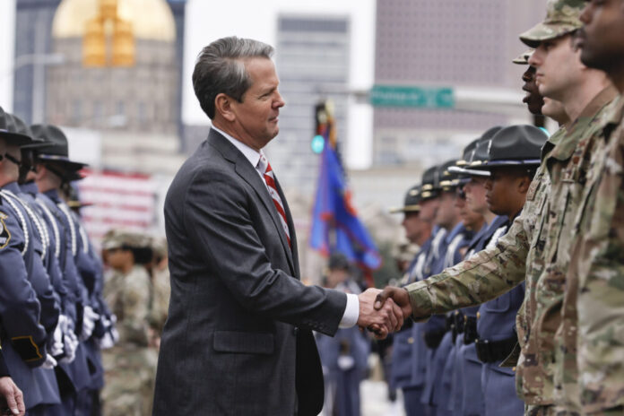 White man in grey suit stands between two lines of military personnel and shakes hands