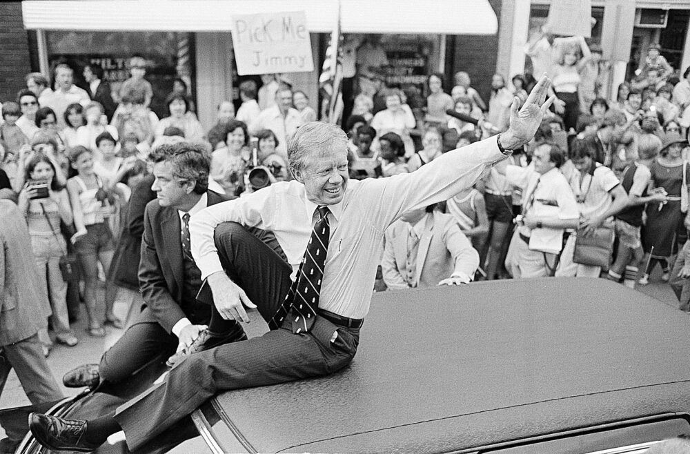 Black and white photo of former President Jimmy Carter sitting on car roof in parade surrounded by crowd