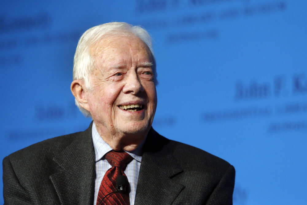 Former President Jimmy Carter in red tie and black jacket in front of blue background