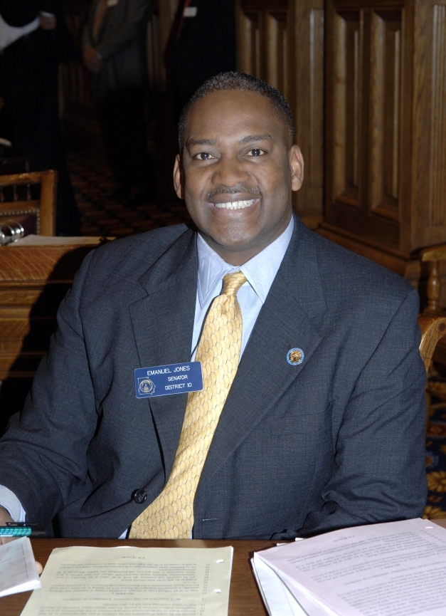 African American man in grey suit with blue shirt and yellow tie sits at brown desk and smiles,
