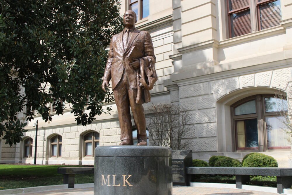 Bronze statue of Martin Luther King standing on black podium that reads "MLK" in front of white building and green trees.
