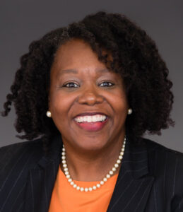 African American woman wearing orange shirt and black blazer poses against grey background