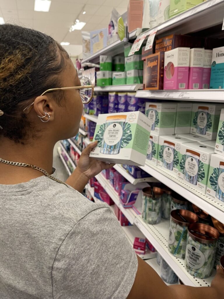 African American woman wearing grey shirt stands in grocery store aisle picking out feminine products.