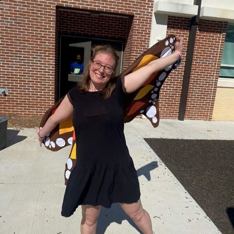 White woman in black dress standing outside in front of brick building poses with butterfly wings. 