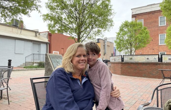 White woman with blonde hair sits in chair hugging young boy with brown hair outside in front of trees and brick building