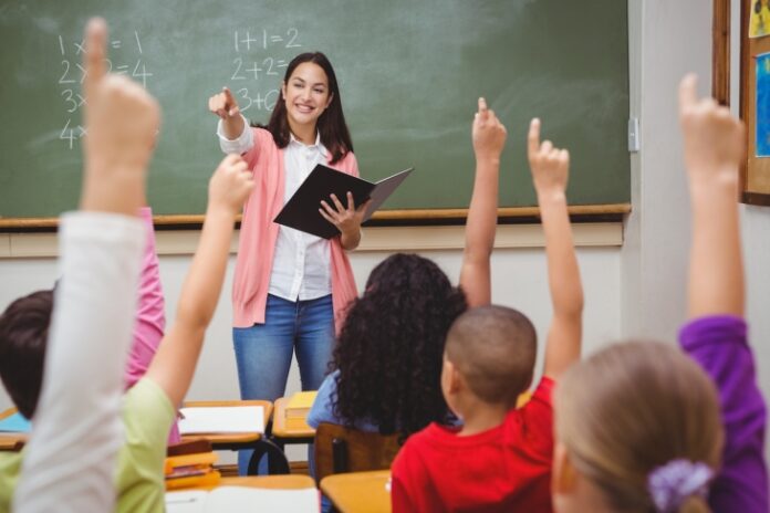 White teacher stands in front of chalkboard with students raising hands