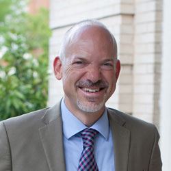 White bald man in beige suit and blue shirt smiling in headshot outdoors