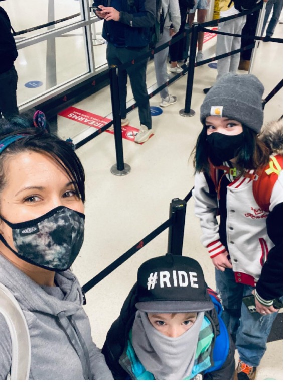 Left to right white mother takes selfie with mask on in airport security line with son and daughter who are both wearing hats and masks