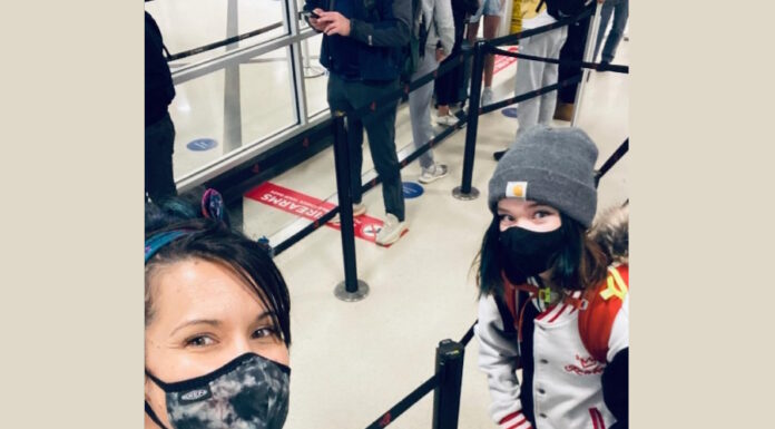Post COVID-19 education in isolation Left to right white mother takes selfie with mask on in airport security line with son and daughter who are both wearing hats and masks