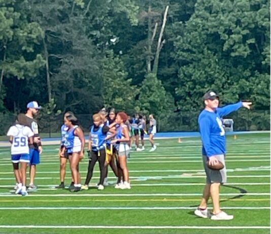 A whole new ball game: Girls flag football touches down in Georgia Women's high school flag football team in blue and white uniforms practice on football field with coach in blue and black with a baseball hat to the right