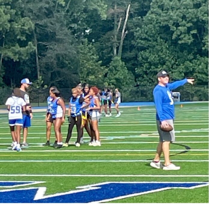Women's high school flag football team in blue and white uniforms practice on football field with coach in blue and black with a baseball hat to the right
