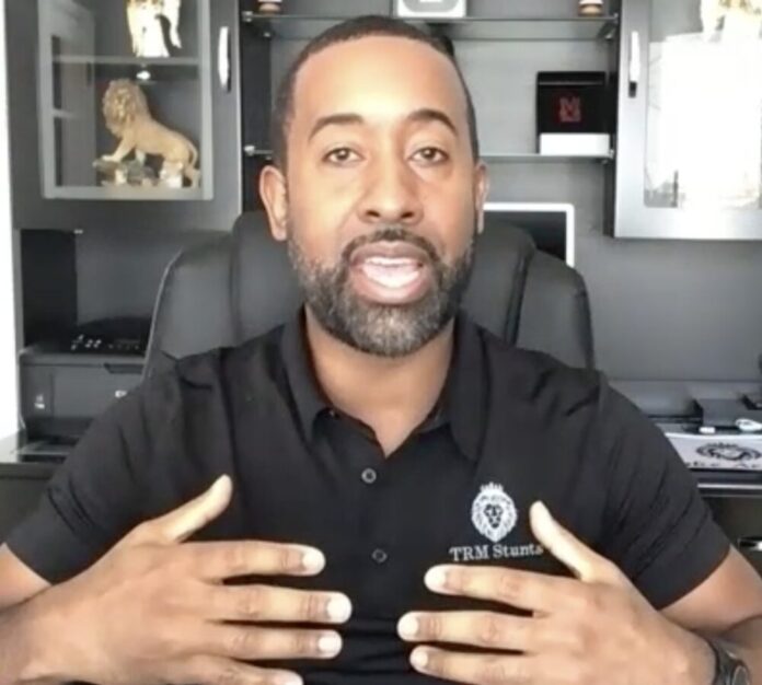 Black man wearing a black polo sitting in office chair at a desk using his hands to illustrate a point while talking.