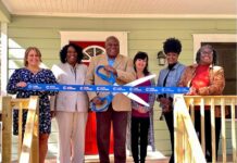 Georgia’s limited maternity homes reach capacity Six people standing on wooden front porch wearing dress clothes and smiling for photo. The black man wearing a tan suit in the center of the photo holds a large pair of blue scissors cutting a blue ribbon outside of a maternity home with light green siding and a red front door.