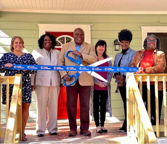 Georgia’s limited maternity homes reach capacity Six people standing on wooden front porch wearing dress clothes and smiling for photo. The black man wearing a tan suit in the center of the photo holds a large pair of blue scissors cutting a blue ribbon outside of a maternity home with light green siding and a red front door.