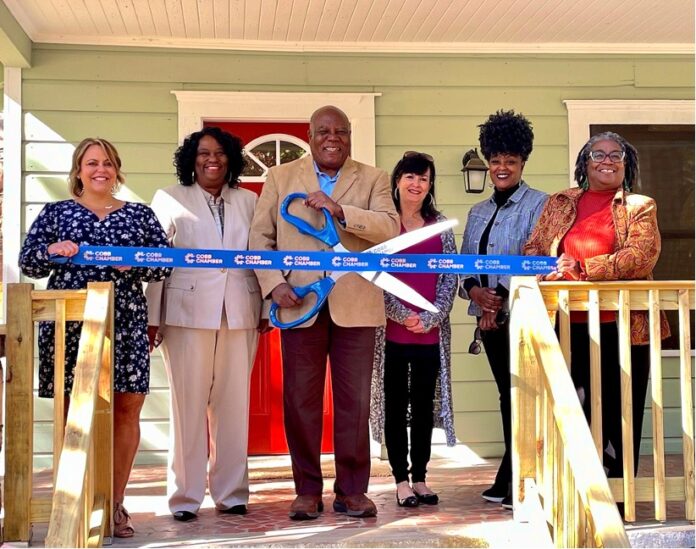 Six people standing on wooden front porch wearing dress clothes and smiling for photo. The black man wearing a tan suit in the center of the photo holds a large pair of blue scissors cutting a blue ribbon outside of a maternity home with light green siding and a red front door.