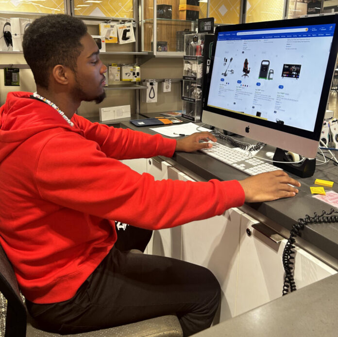 A Black man in a red hoodie sits at a Macbook scrolling through Walmart's website looking at Black Friday deals.