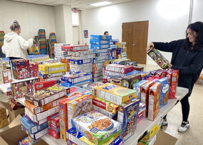 Three volunteers stand around a table piled with dozens of boxed food items.