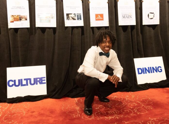 Atlanta student homelessness: Young Black man with black dreads in formal wear with black bow tie crouches on orange carpet in front of black curtain with several hanging paper signs.