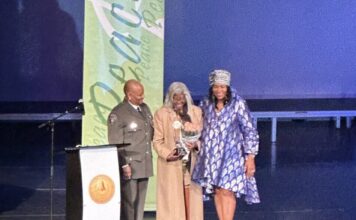Cobb NAACP presents annual “Living the Dream” award on MLK Day MLK award ceremony: Three Black women stand together on a stage behind a podium. On the left, the woman wears a police uniform. In the center, the woman wears a long coat and holds a trophy and bouquet of flowers. The woman on the right wears a blue dress.