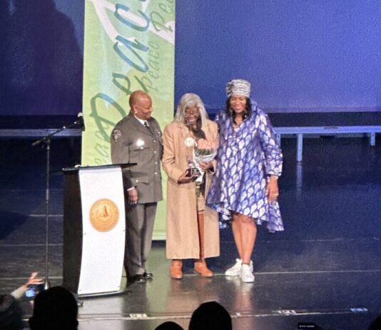 Cobb NAACP presents annual “Living the Dream” award on MLK Day MLK award ceremony: Three Black women stand together on a stage behind a podium. On the left, the woman wears a police uniform. In the center, the woman wears a long coat and holds a trophy and bouquet of flowers. The woman on the right wears a blue dress.