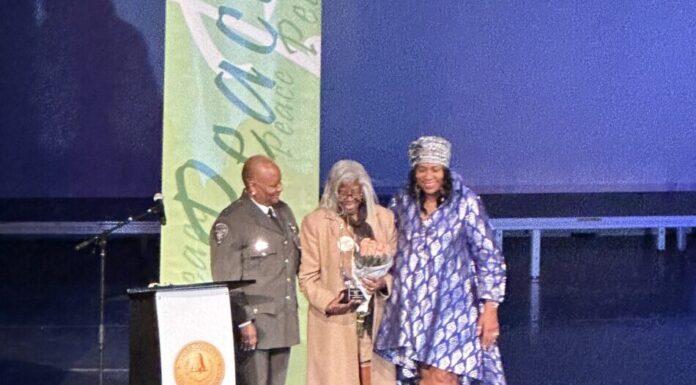 Cobb NAACP presents annual “Living the Dream” award on MLK Day MLK award ceremony: Three Black women stand together on a stage behind a podium. On the left, the woman wears a police uniform. In the center, the woman wears a long coat and holds a trophy and bouquet of flowers. The woman on the right wears a blue dress.