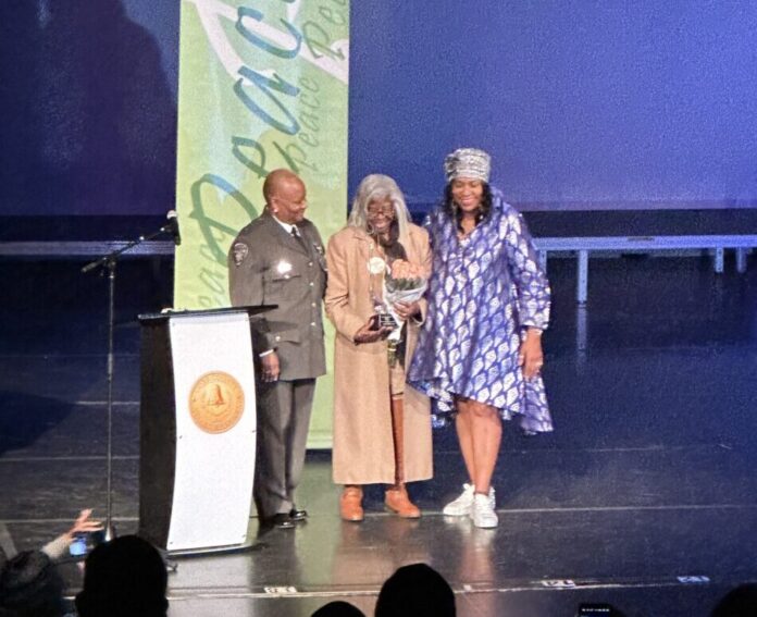 MLK award ceremony: Three Black women stand together on a stage behind a podium. On the left, the woman wears a police uniform. In the center, the woman wears a long coat and holds a trophy and bouquet of flowers. The woman on the right wears a blue dress.