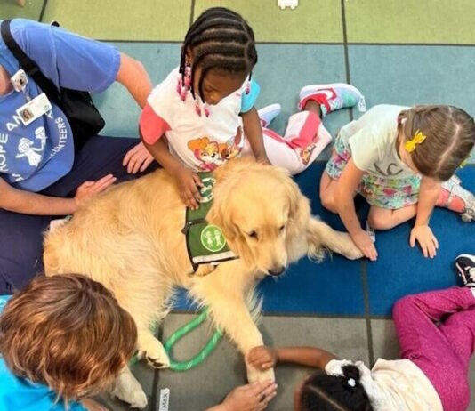 Everyday Hero: Reggie lends a helping paw GA Everyday Heroes: Helping Paw - Several; young children and an adult sitting on floor in circle around large, golden-furred dog wearing a green vest