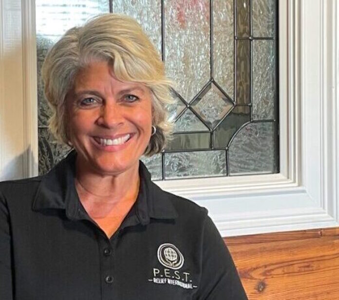 GA Everyday Heroes: Headshot Andrea Hancock middle-aged woman with short silver hair in black shirt with P.E.S.T. round logo in white, in front of leaded glass window of red brick building