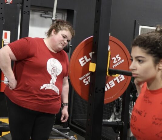 Champion female powerlifter coaches next generation to victory Powerlifter Becci Holcomb wears a red "Pretty Strong Powerlifting" t-shirt. She is watching a girl with a red shirt lifting weights and coaching her. They are both standing in a gym with black walls and floors.