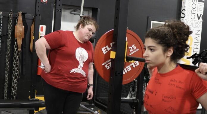 Champion female powerlifter coaches next generation to victory Powerlifter Becci Holcomb wears a red "Pretty Strong Powerlifting" t-shirt. She is watching a girl with a red shirt lifting weights and coaching her. They are both standing in a gym with black walls and floors.