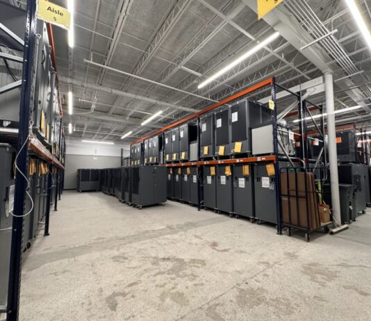 Georgia voting machine overhaul raises concerns for upcoming elections Wide shot interior warehouse with concrete floors and fluorescent lights on ceiling. There are tall rows of shelves containing dozens of large Dominion voting machines.