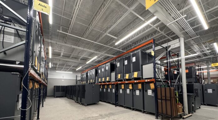 Georgia voting machine overhaul raises concerns for upcoming elections Wide shot interior warehouse with concrete floors and fluorescent lights on ceiling. There are tall rows of shelves containing dozens of large Dominion voting machines.