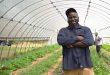 Community is the strongest ally for Black farmers’ mental health struggles A Black man wearing a baseball cap and navy blue button-up shirt stands with his arms crossed and smiles at the camera. He is standing in a rounded glass enclosure with rows of crops growing behind him.