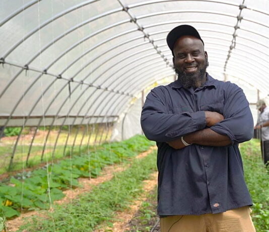 Community is the strongest ally for Black farmers’ mental health struggles A Black man wearing a baseball cap and navy blue button-up shirt stands with his arms crossed and smiles at the camera. He is standing in a rounded glass enclosure with rows of crops growing behind him.