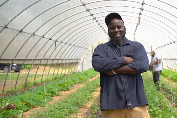 A Black man wearing a baseball cap and navy blue button-up shirt stands with his arms crossed and smiles at the camera. He is standing in a rounded glass enclosure with rows of crops growing behind him.