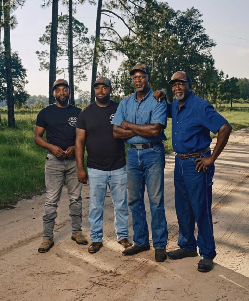 Four Black men stand together on a dirt road in front of a patch of trees. Two men on the left both wear jeans and black Berry Farms shirts. Two men on the right wear denim button-ups and matching jeans. All four men wear matching Berry Farms baseball hats.