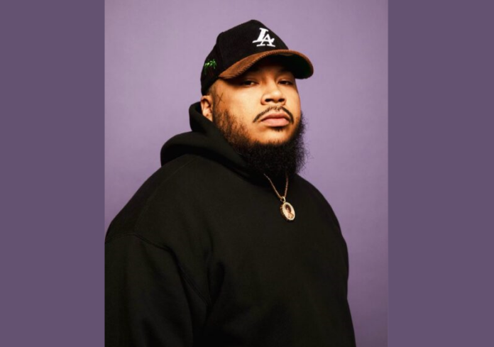 Headshot of Kali Cunningham, a young adult male with Los Angeles Dodgers hat and black beard wearing black hooded sweatshirt and a gold chain against a plain purple background.