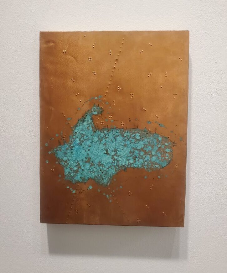 A rectangular hammered copper plate with engraved braille detailing and a turquoise shape resembling a pond hangs on a white wall.