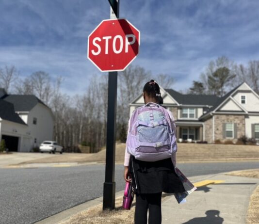 Georgia GOP pushes to pass school voucher bill this year A young Black girl wearing a school uniform and a purple backpack stands on the sidewalk in front of a stop sign. She is photographed from behind.