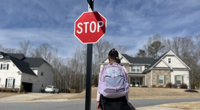 Georgia GOP pushes to pass school voucher bill this year A young Black girl wearing a school uniform and a purple backpack stands on the sidewalk in front of a stop sign. She is photographed from behind.