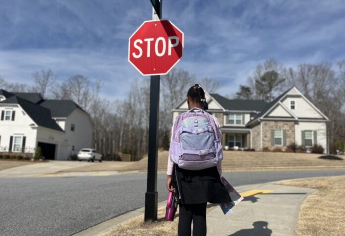 A young Black girl wearing a school uniform and a purple backpack stands on the sidewalk in front of a stop sign. She is photographed from behind.