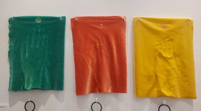Visitors break all the rules at hands-on art exhibit Three pieces of velvet embroidered with hands are displayed on a white wall. From left to right, the pieces are green, red, and yellow.