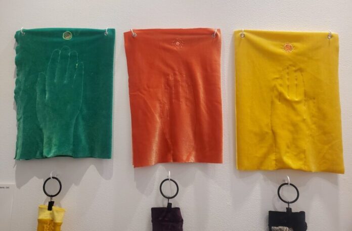 Three pieces of velvet embroidered with hands are displayed on a white wall. From left to right, the pieces are green, red, and yellow.
