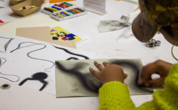 High Museum of Art offers free admission for all A woman sits at a table surrounded by oil pastels. She wears a hat and green sweater and is drawing large, swirling black lines with an oil pastel on paper.